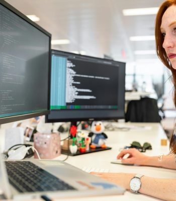 A focused female software engineer coding on dual monitors in a modern office.